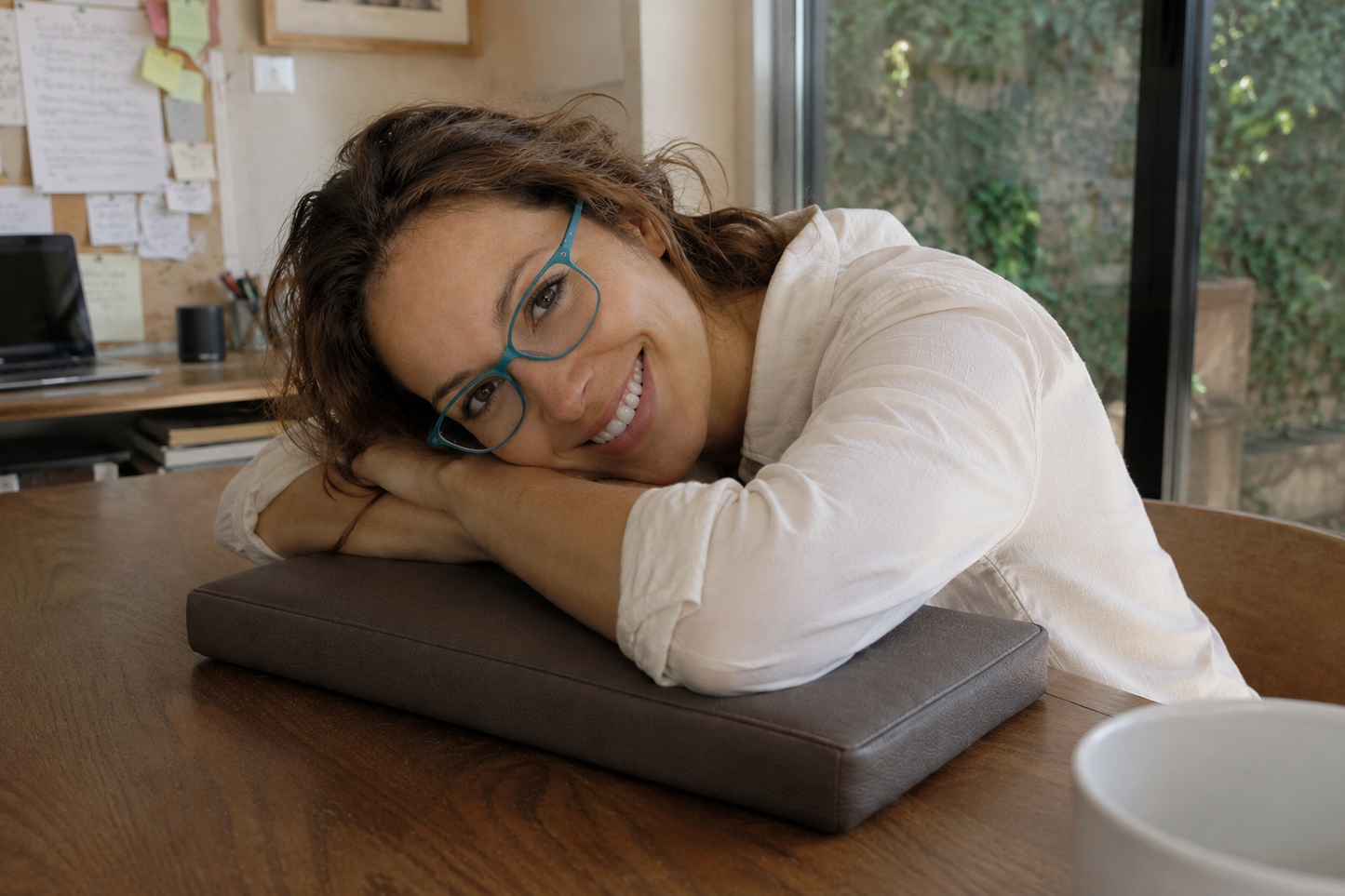 A woman with brown hair and blue glasses rests her head on a dark brown rectangular cushion on a wooden table in an office setting.